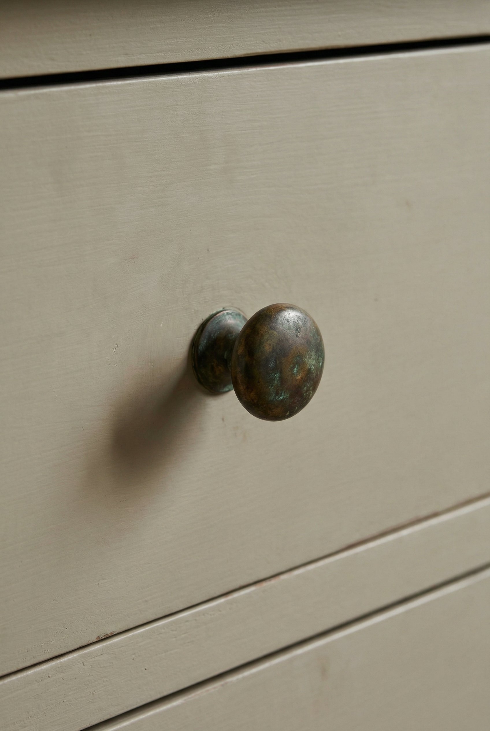 Photorealistic interior photo. Close up of an aged, unlacquered brass cabinet knob on a putty-painted flat-panel drawer, heavy natural patina, soft shadow. Editorial photography style, no people visib