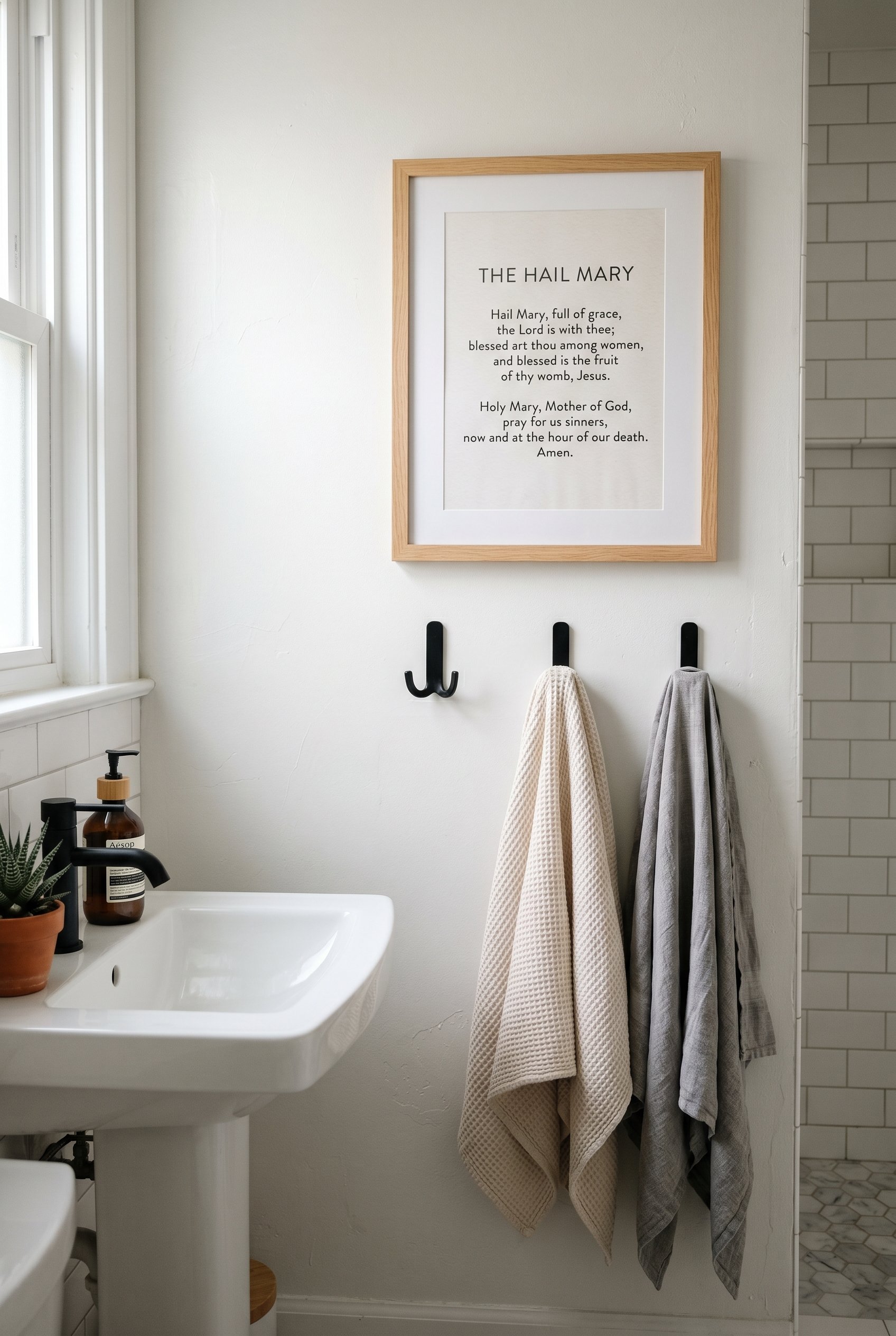 Photorealistic interior photo. A clean, modern typography print of a Catholic prayer in a simple oak frame hanging above matte black towel hooks in a bright, modern bathroom. Editorial photography sty