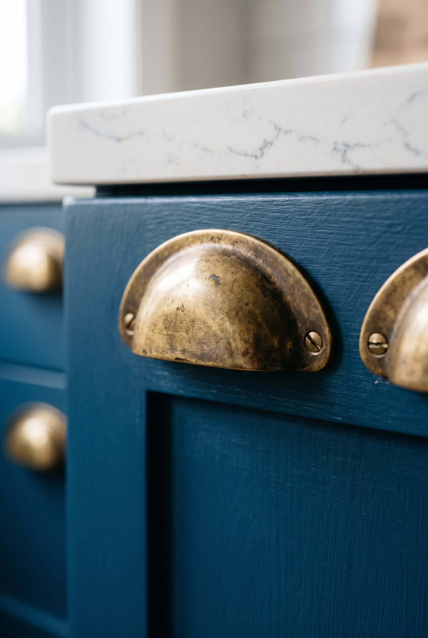 Photorealistic interior photo. Close up macro shot of unlacquered brass cup pulls on a French blue kitchen drawer. Soft shadows, editorial photography style.