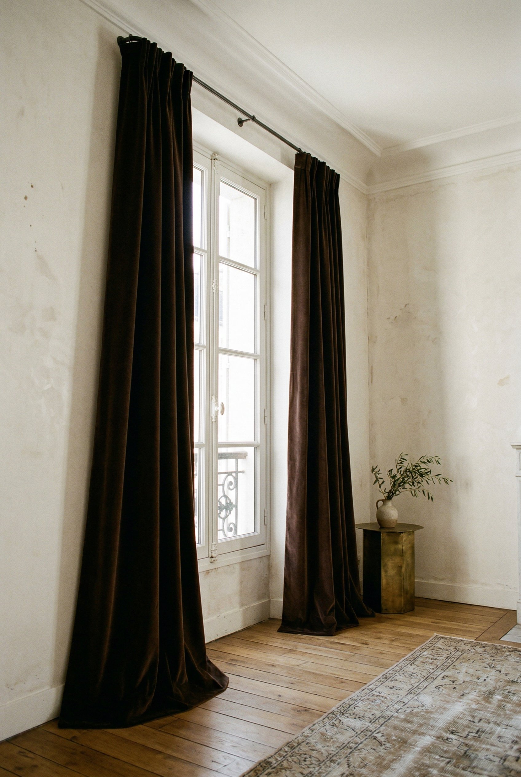 Photorealistic interior photo. Heavy, textured velvet drapes in deep chocolate brown, hung high on a simple French return rod, lightly kissing the floor, bright window light, lower angle. Editorial ph