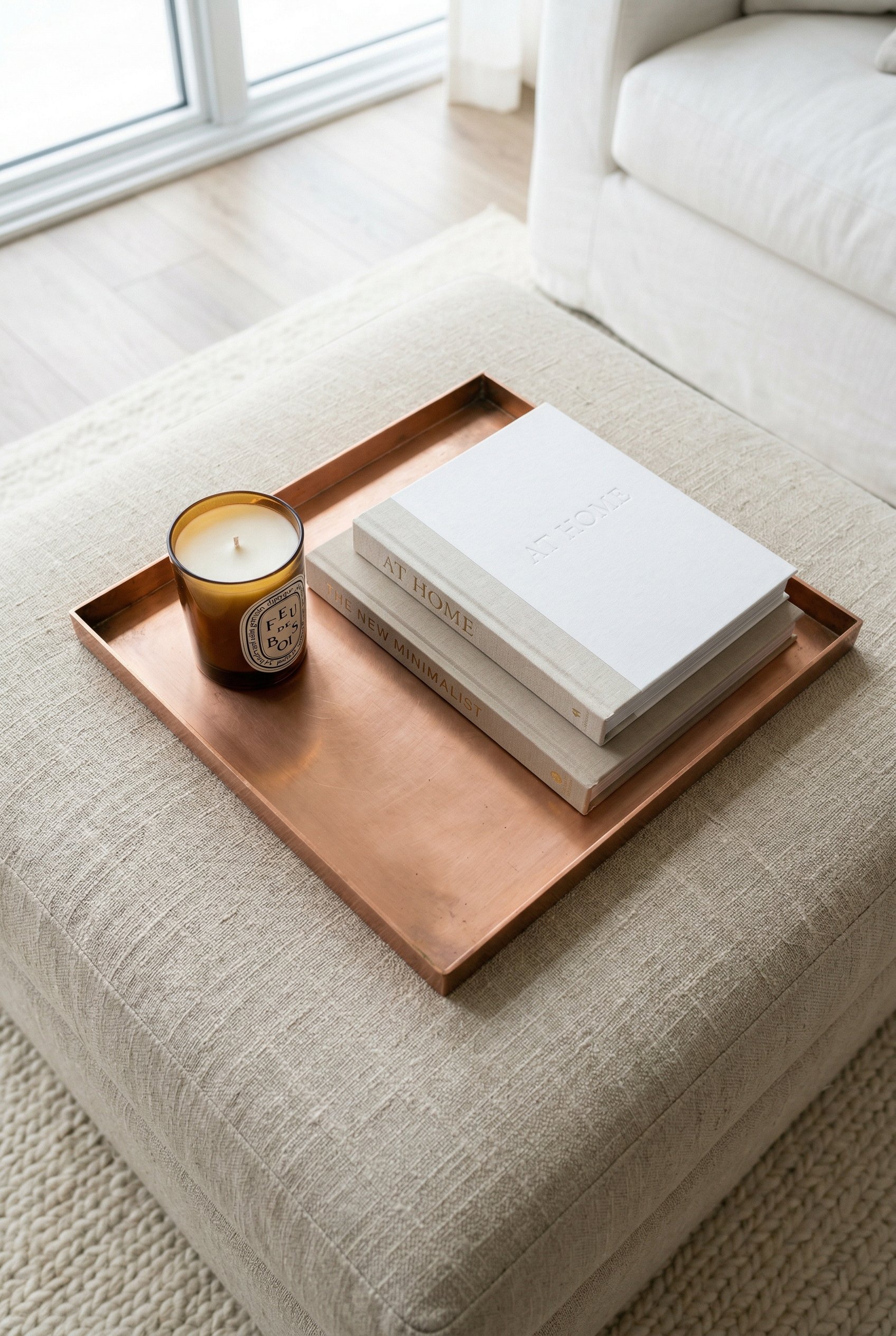 Photorealistic interior photo. Minimalist flat copper tray resting on a beige linen ottoman, holding a luxury candle and two design books, bright diffused daylight, top-down angle. Editorial photograp