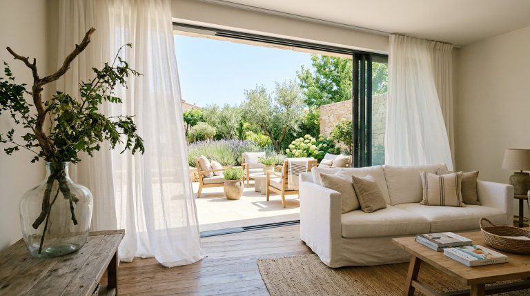Photorealistic interior photo of a summer home interior blurring indoor and outdoor living. Wide open sliding glass doors leading to a bright patio. Inside, a crisp white slipcovered sofa, a large for