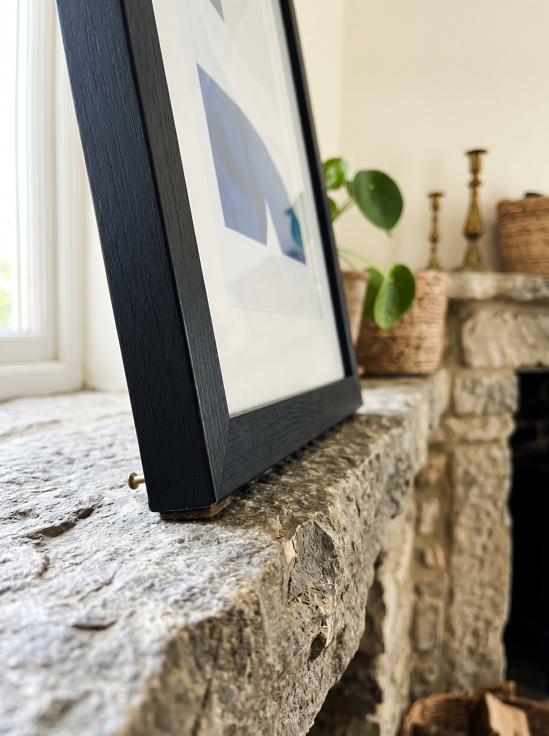 Photorealistic interior photo. Close-up detail of the bottom corner of a black picture frame leaning securely on a stone mantel ledge. Focus on the crisp intersection of frame and stone. Bright, clear