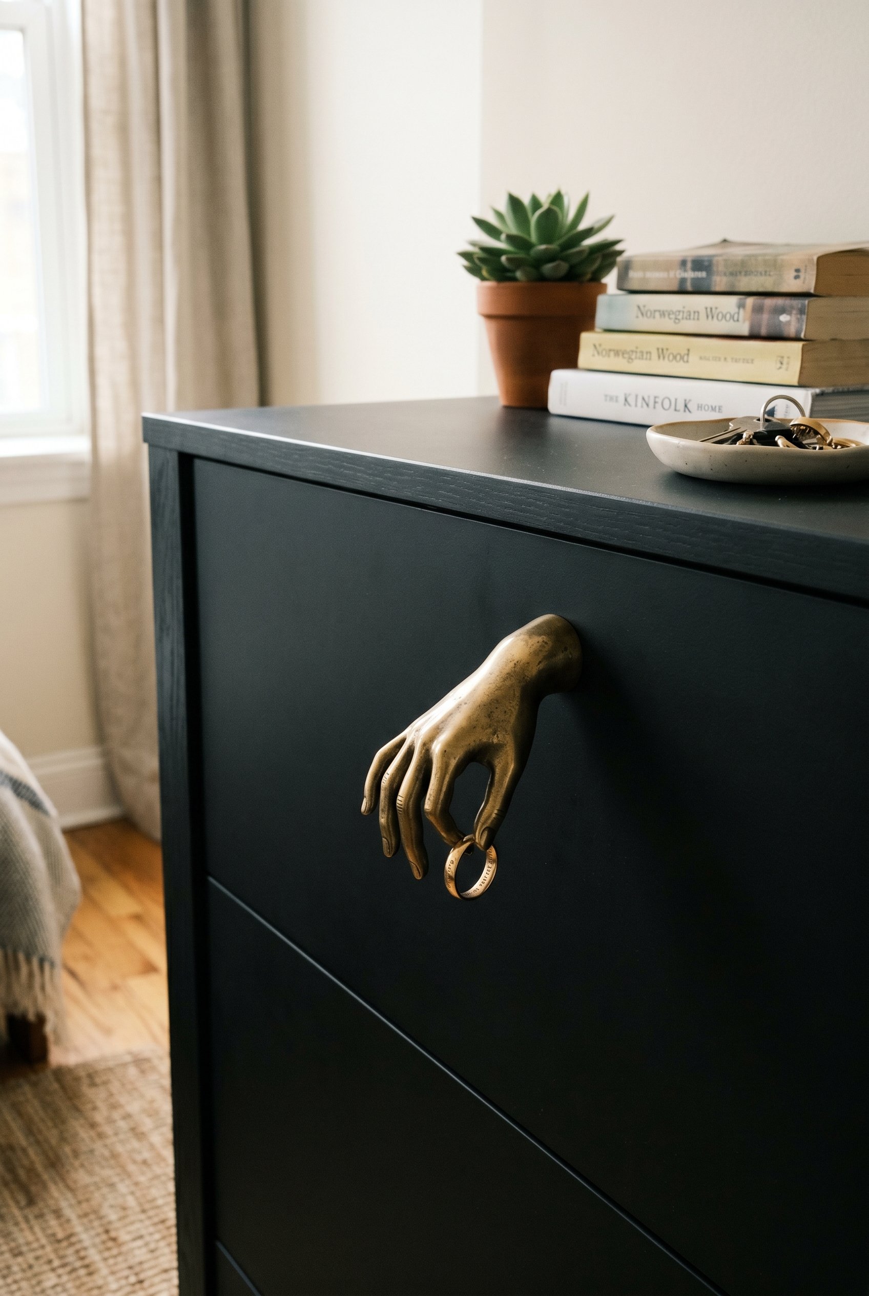 Photorealistic interior photo. Close up of a sleek matte black dresser drawer featuring a solid brass drawer pull shaped like a surrealist human hand holding a ring, sharp focus. Editorial photography